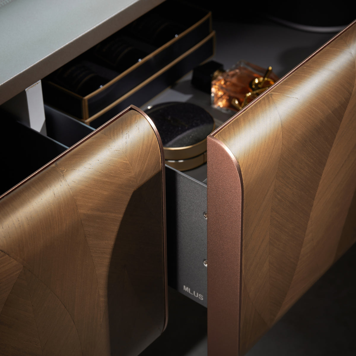 Close-up of a luxury vanity drawer with a geometric chevron wood grain finish in a warm bronze-brown tone, shown partially open with cosmetics inside.