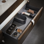 An open bathroom vanity drawer with a dark gray interior organizer, brown wood-look exterior, and visible toiletries including a perfume bottle, compact, and cylindrical container.