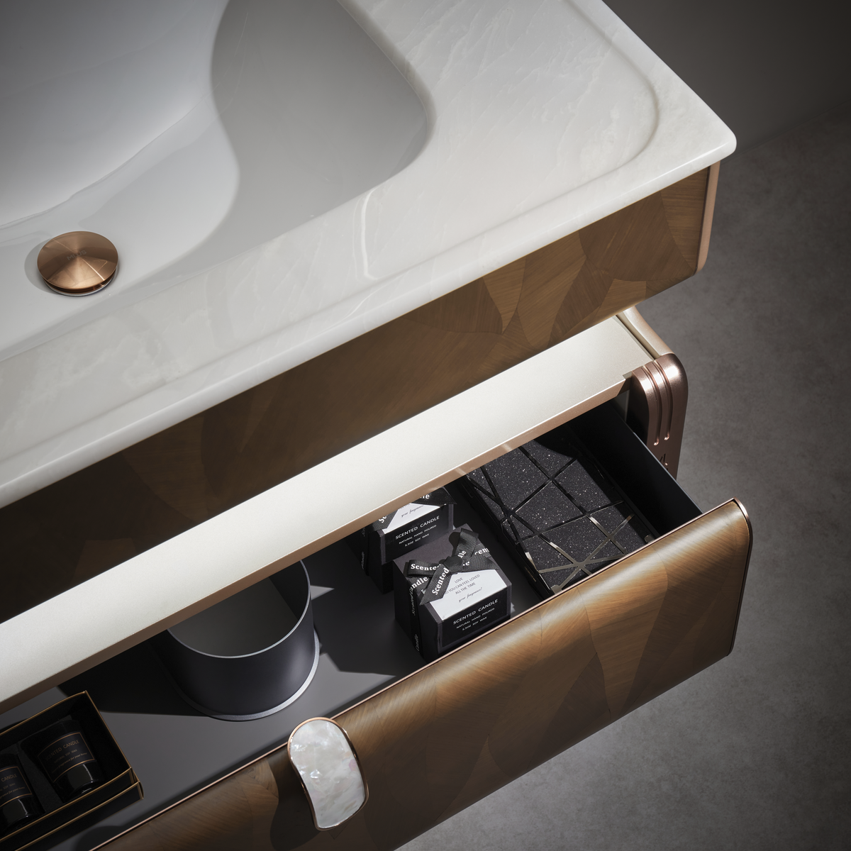 Close-up of a modern bathroom vanity with a white integrated sink, geometric bronze-brown cabinet front, and an open lower drawer showing toiletries and black bath mats on a dark surface.